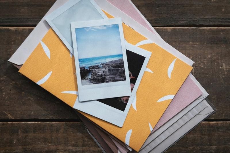 Two instant photos, one of a rocky shoreline, on a stack of colored envelopes on a wooden table