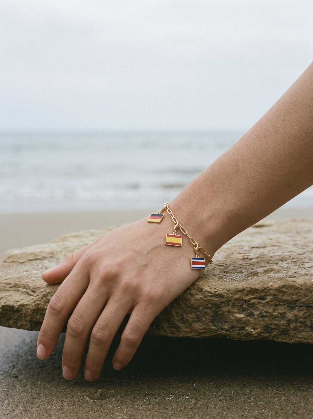 Gold travel charm bracelet with three flag charms on a wrist at the coast